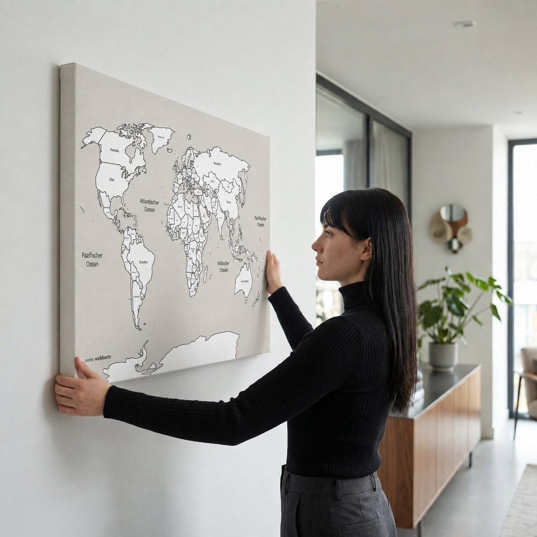 Eine Frau richtet eine beige Weltkarte mit Ländernamen auf Keilrahmen an einer weißen Wand aus.